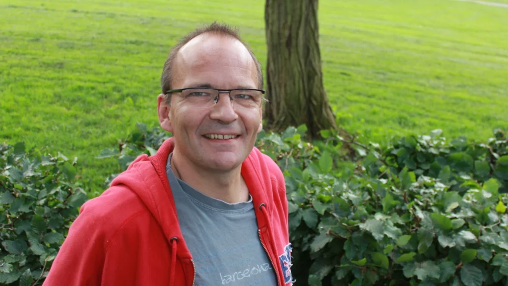 Frederik Hostrup smiler udendørs i en park med grøn natur i baggrunden.
