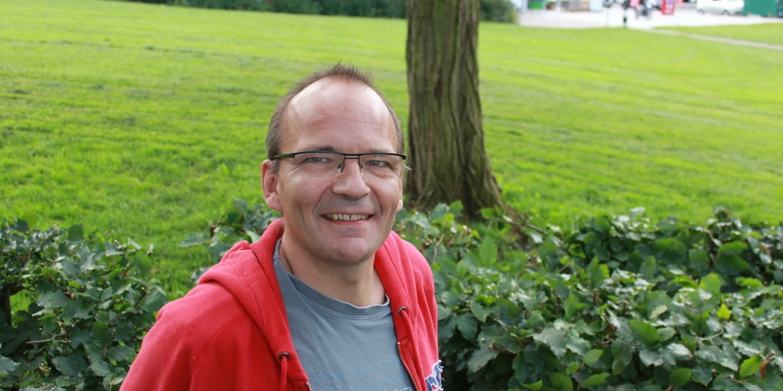Frederik Hostrup smiler udendørs i en park med grøn natur i baggrunden.