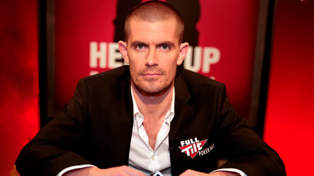 Gus Hansen sidder ved et pokerbord i jakkesæt under en pokersending og kigger direkte mod kameraet.