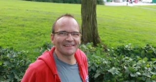 Frederik Hostrup smiler udendørs i en park med grøn natur i baggrunden.